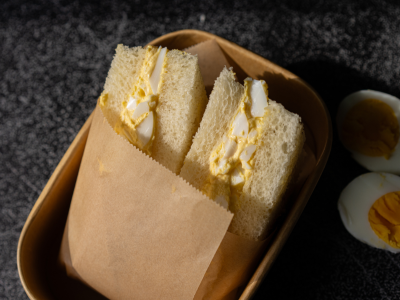 Two egg sandwiches in a paper wrapper inside a brown tray, with a cut boiled egg beside.
